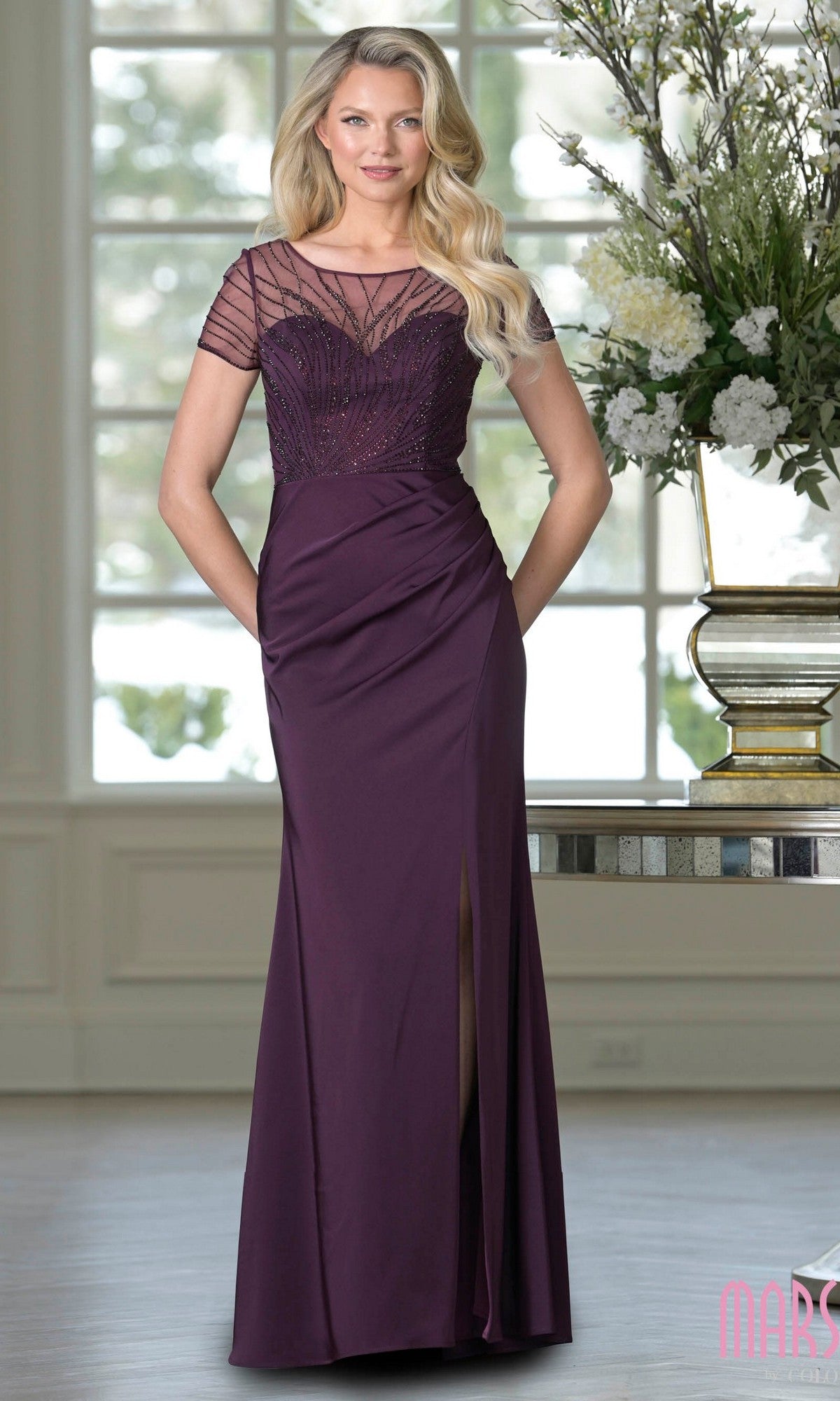 A woman stands indoors near a vase of white flowers wearing the Marsoni M350, a long formal eggplant dress with a beaded bodice, short sleeves, sheath skirt, and slit.